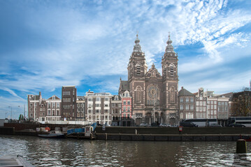 Fototapeta premium Historic Amsterdam cityscape with the Basilica of Saint Nicholas prominently featured. The scene is under a bright blue sky with scattered clouds