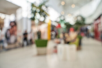 Obraz premium Blurred shopping mall interior with people and plants beneath bright skylight in busy retail setting.