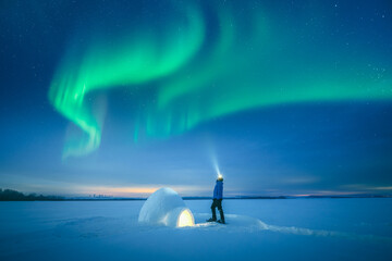 Polar survival concept with man using flashlight near illuminated snow igloo in open winter field, aurora borealis, arctic night scene, polar lights, northern lights © Ivan Kmit