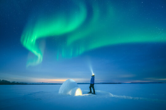 Polar survival concept with man using flashlight near illuminated snow igloo in open winter field, aurora borealis, arctic night scene, polar lights, northern lights