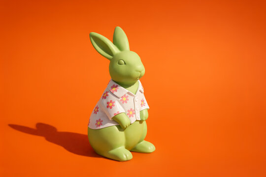 Green rabbit figurine wearing floral shirt on bright orange background. Playful character, bold color contrast, minimal composition, decorative seasonal still life with cheerful mood. Easter concept. 