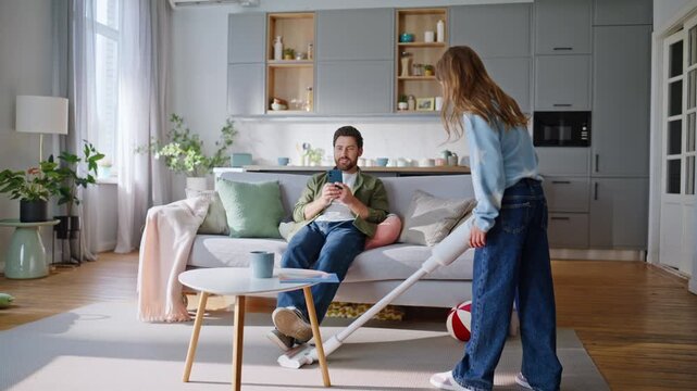 Helpful girl cleaning apartment vacuum cleaner. Father relaxing couch