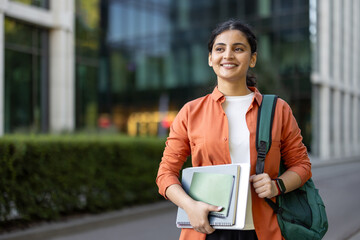 Obraz premium Young happy female student carrying a backpack and notebooks, smiling and looking ahead while walking confidently on a university campus, embodying education, growth, and optimism for the future