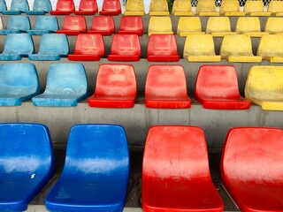 Naklejka premium Rows of colorful plastic stadium seats in blue, red, and yellow at an outdoor sports arena, creating a vibrant geometric pattern in an empty spectator area