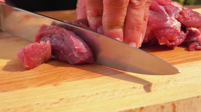 Knife cutting fresh raw pork meat into small pieces on a wooden board. Cooking process in the kitchen. Preparation of ingredients for meal.