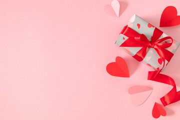 Festive arrangement for Valentine's Day: a gift box with a red ribbon and paper hearts on a pastel pink background. Valentine's Day gift. Top view, flat lay.