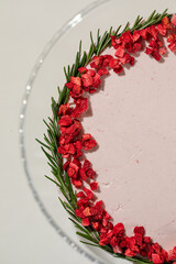 Elegant pink cake decorated with red berries and rosemary served on glass stand with patterned background