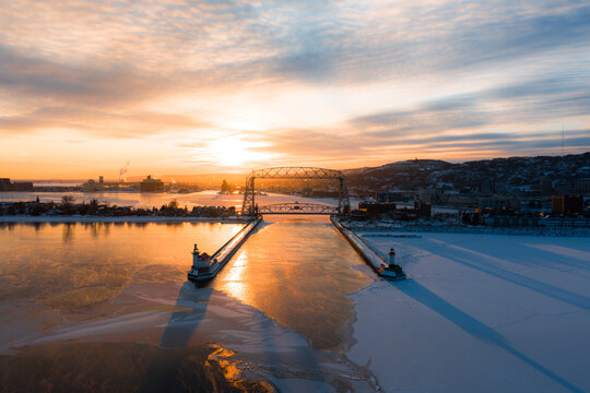 A wide aerial perspective of Duluth's Canal Park, the Aerial Lift Bridge, and the lighthouses under a brilliant winter sunrise.