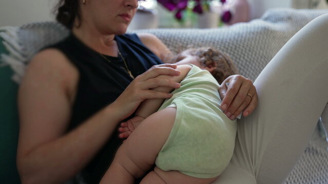 Mother breastfeeding curly-haired toddler while reclining on sofa, close-up of baby in green onesie lying across lap and nursing in quiet intimate home setting