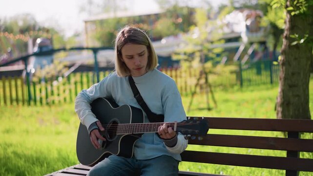 young white male guitarist busking in park, bright green lawn with distant carousel, strumming upbeat chords for passerby, warm daylight, casual denim, smiling audience, lively weekend atmosphere,