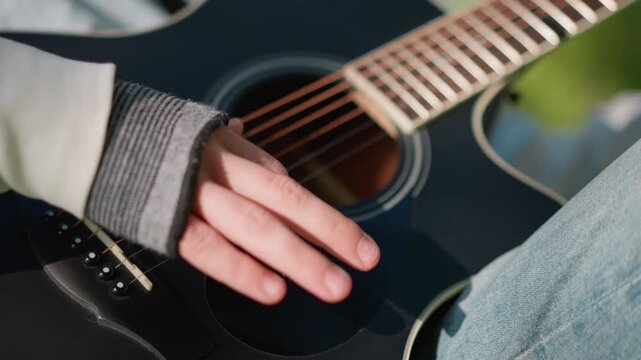 closeup acoustic guitar hand strumming, casual guitarist wearing grey sleeve and denim, fingers brushing strings over black guitar body, sunny park background, student musician practicing chord