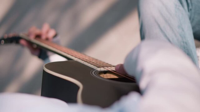 overhead closeup guitarist playing acoustic, focus on fretting hand, denim jeans, blonde hair fringe, sunlight stripe, bench setting, upbeat fingerpicking tutorial, busking mood, subtle shadows, warm