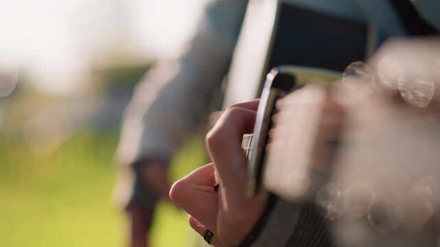 Closeup hands strumming acoustic guitar, outdoor park session with warm sunlight and creamy bokeh, capo clamped near nut, ring on finger, casual jacket sleeve, intimate fingerstyle playing, shallow