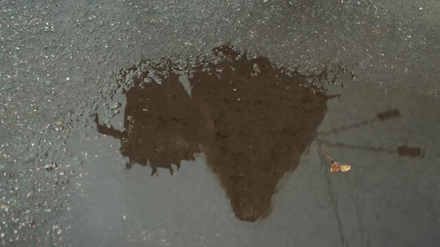 wet asphalt puddle with single leaf, close aerial view of water spreading across textured pavement dark irregular stain expands into reflective pool, subtle ripples mirror gray sky, damp pebbled