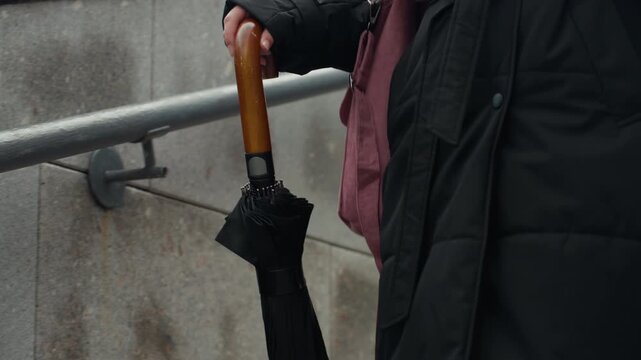 closeup woman holding umbrella handle stairs, long black coat and pink bag visible, steady grip on wooden curved handle, descending stone steps beside metal handrail, overcast light and muted palette,