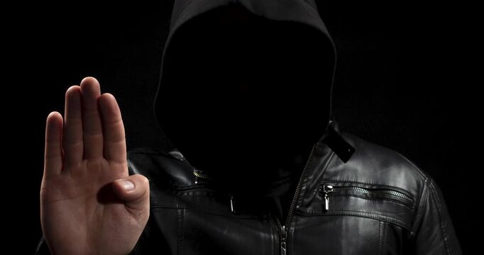 a zoom out of a hooded hacker man in the shadows with his right hand making a stop signal and a black background