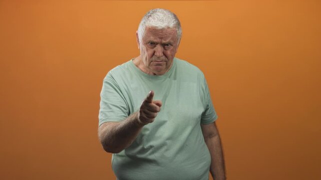 Senior man points finger to camera in studio with orange backdrop, wearing mint t shirt and furrowed brow; reprimand warning.