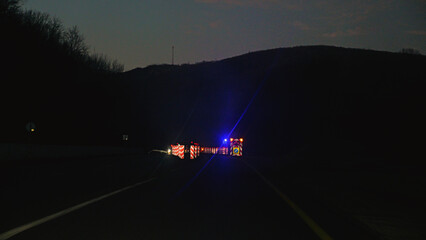 View from a car of a highway road block at dusk. Flashing blue emergency lights and reflective barriers signal a warning and detour. Dark, moody, and cautious atmosphere with lens flare © VeNN