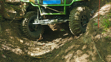 Fototapeta premium Powerful off-road vehicle with large muddy tires and visible suspension struggling through a deep dirt trench in a forest. Extreme sport competition with dappled sunlight