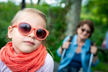  Cute Toddler Girl in Orange Sunglasses and Lace Scarf Portrait with Blurred Hiking Grandma on Forest Trail