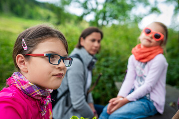 Serious Young Girl in Red Frame Glasses and Pink Plaid Scarf Portrait with Blurred Mom and Sister in Forest Nature