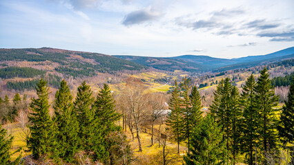 Obraz premium A wide view of Swieradow Zdroj in Poland shows rolling hills and trees under a blue sky. The landscape has patches of grass and areas of bare trees. Mountains rise in the distance.