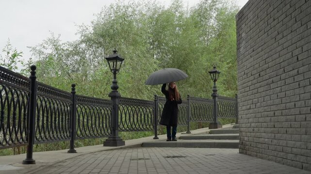 man walking under black umbrella along cobblestone walkway by iron fence and vintage lampposts, overcast sky, solitary mood, slow measured pace, coat and subtle drizzle, scattered leaves, cinematic