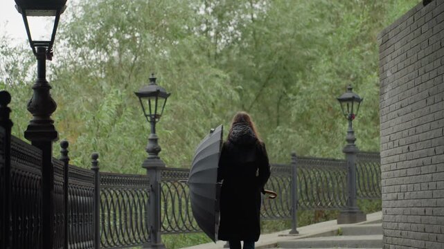 person walking with umbrella along fence and vintage lamps, passing brick wall beneath overcast sky quiet stroll through green trees creates moody reflective atmosphere with raindrops and shadowed