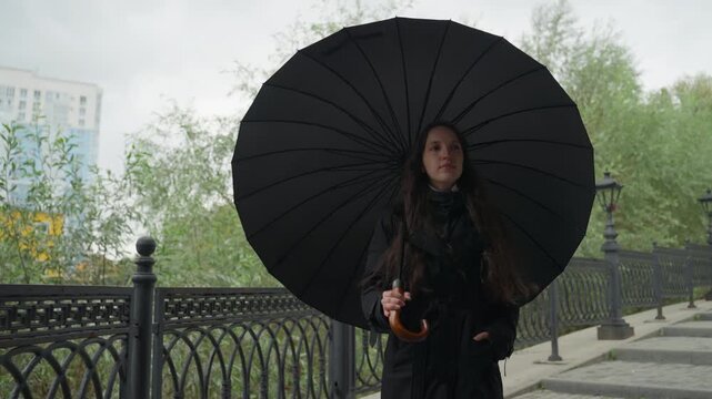 woman with black umbrella walking along riverside walkway past ornate railing and lampposts under overcast sky with raindrops, contemplative mood, elegant coat and large parasol framed as silhouette