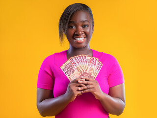 A woman with a bright smile holds a fan of 200 Ghanaian cedi notes against a vibrant yellow background. She wears a pink shirt, radiating joy and financial success in this studio-style portrait.
