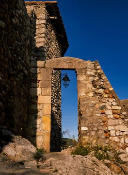 Historic stone archway with a vintage lantern in Ardeche, France. Sunlight illuminating ancient masonry ruins against a deep blue sky.