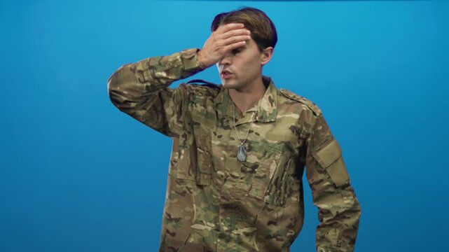 Man in military uniform with dog tags touching forehead with hand and pressing hand to chest in studio; fatigue duty.