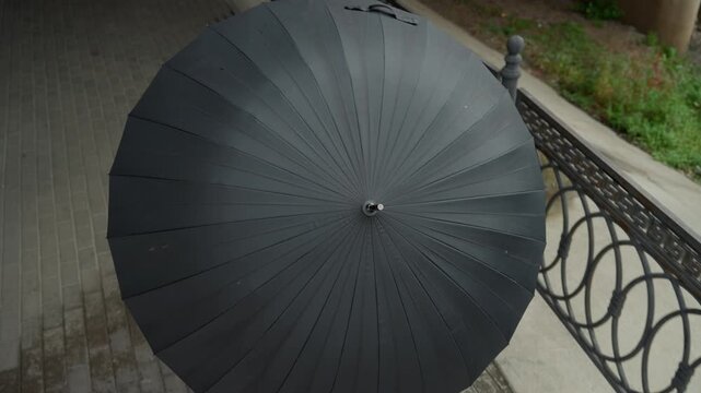 topdown black umbrella over pavement, iron railing and puddles visible, subtle raindrops on textured canopy, radial pattern and knob detail, moody overcast light, cinematic close perspective