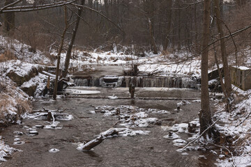 Wodospad na niewielkiej rzece. Jest zima. Brzegi pokryte są warstwą śniegu. Rzekę miejscami pokrywa lód. Wzburzona woda na wodospadzie przepływa pomiędzy lodowymi czapami. © boguslavus