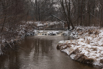 Wodospad na niewielkiej rzece. Jest zima. Brzegi pokryte są warstwą śniegu. Rzekę miejscami pokrywa lód. Wzburzona woda na wodospadzie przepływa pomiędzy lodowymi czapami. © boguslavus