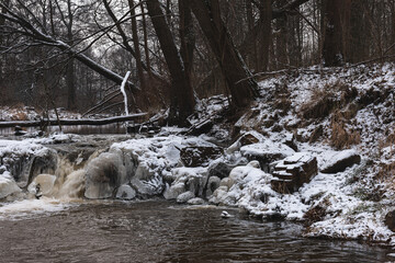 Wodospad na niewielkiej rzece. Jest zima. Brzegi pokryte są warstwą śniegu. Rzekę miejscami pokrywa lód. Wzburzona woda na wodospadzie przepływa pomiędzy lodowymi czapami. © boguslavus