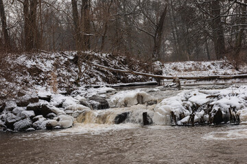 Wodospad na niewielkiej rzece. Jest zima. Brzegi pokryte są warstwą śniegu. Rzekę miejscami pokrywa lód. Wzburzona woda na wodospadzie przepływa pomiędzy lodowymi czapami. © boguslavus