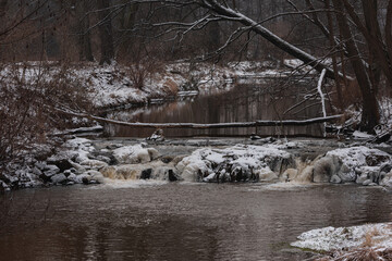 Wodospad na niewielkiej rzece. Jest zima. Brzegi pokryte są warstwą śniegu. Rzekę miejscami pokrywa lód. Wzburzona woda na wodospadzie przepływa pomiędzy lodowymi czapami. © boguslavus
