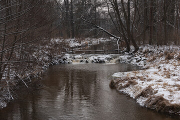 Wodospad na niewielkiej rzece. Jest zima. Brzegi pokryte są warstwą śniegu. Rzekę miejscami pokrywa lód. Wzburzona woda na wodospadzie przepływa pomiędzy lodowymi czapami. © boguslavus