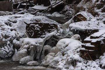 Wodospad na niewielkiej rzece. Jest zima. Brzegi pokryte są warstwą śniegu. Rzekę miejscami pokrywa lód. Wzburzona woda na wodospadzie przepływa pomiędzy lodowymi czapami. © boguslavus