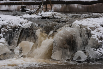 Wodospad na niewielkiej rzece. Jest zima. Brzegi pokryte są warstwą śniegu. Rzekę miejscami pokrywa lód. Wzburzona woda na wodospadzie przepływa pomiędzy lodowymi czapami. © boguslavus