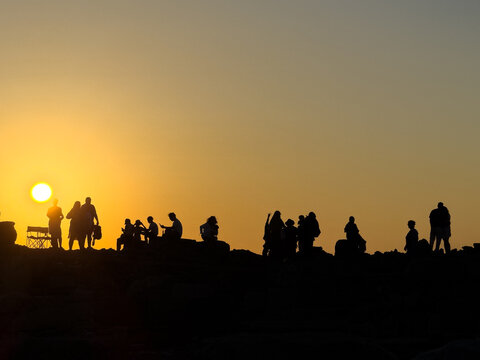 "Silhouettes of a crowd of tourists watching a golden sunset on a rocky landscape"
