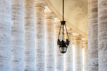 Naklejka premium Visitors walk through the colonnade at St Peters Square in Vatican. The tall columns create a grand space under the ceiling. A light fixture hangs down, adding to the design of the area.