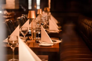 Fotobehang Horeca Elegant restaurant interior with a long table set for dinner, neatly folded white napkins, wine glasses, plates and cutlery, warm ambient lighting from small table lamps, cozy and intimate atmosphere,  © Валентина Руденька
