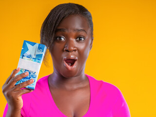 A woman looks wide-eyed and shocked while holding a thick stack of blue 5 Ghana Cedis banknotes. Set against a bold yellow background, her vibrant pink shirt makes the Ghanaian currency pop.