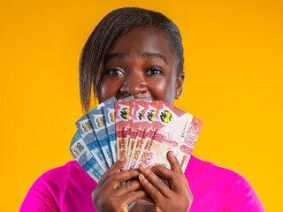 A woman with a dark complexion holds a fan of Ghanaian Cedi notes (GH₵100 and GH₵200) against a bright yellow background. She wears a pink top, her eyes wide with excitement behind the currency.