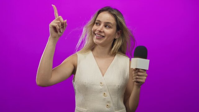 Woman holding microphone and points finger up in magenta studio, smiling at camera and gesturing with raised hand; confidence reporting.