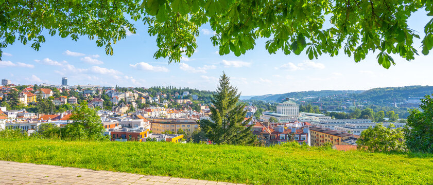 The scenery shows the Podol district in Prague with buildings and trees. People can be seen enjoying the sunny day. The background includes hills and a clear sky.