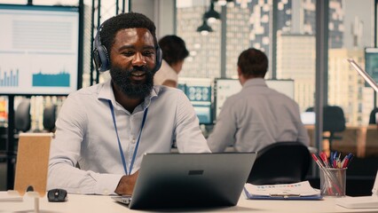 Fototapeta premium Black professional attending video conference with headphones in modern office. Business work scene showing charts and data analysis during online briefing, transparency and communication.