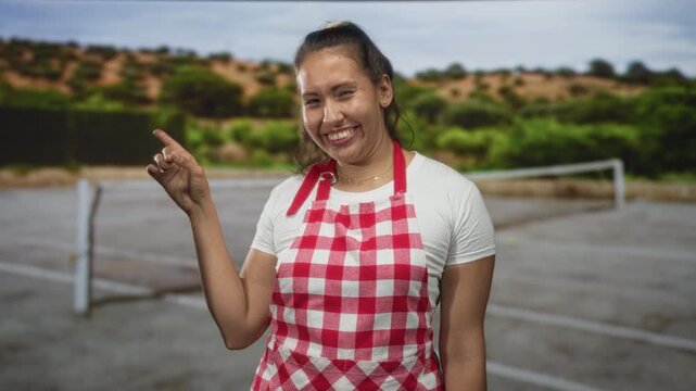 Woman cook wearing red gingham apron points finger on street tennis court while smiling and winking with relaxed stance; playful joy.